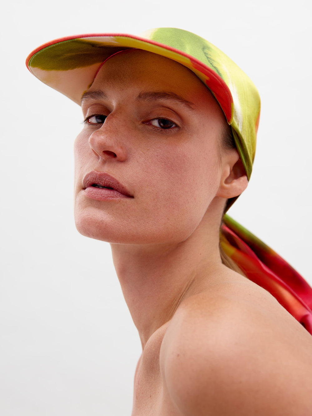A person wearing the Tesia Cap Spring Blooms, featuring a draped back extension and multicolored design, looks straight at the camera against a plain background. Don't miss out—pre-order now! Ships by February 15th, 2025.