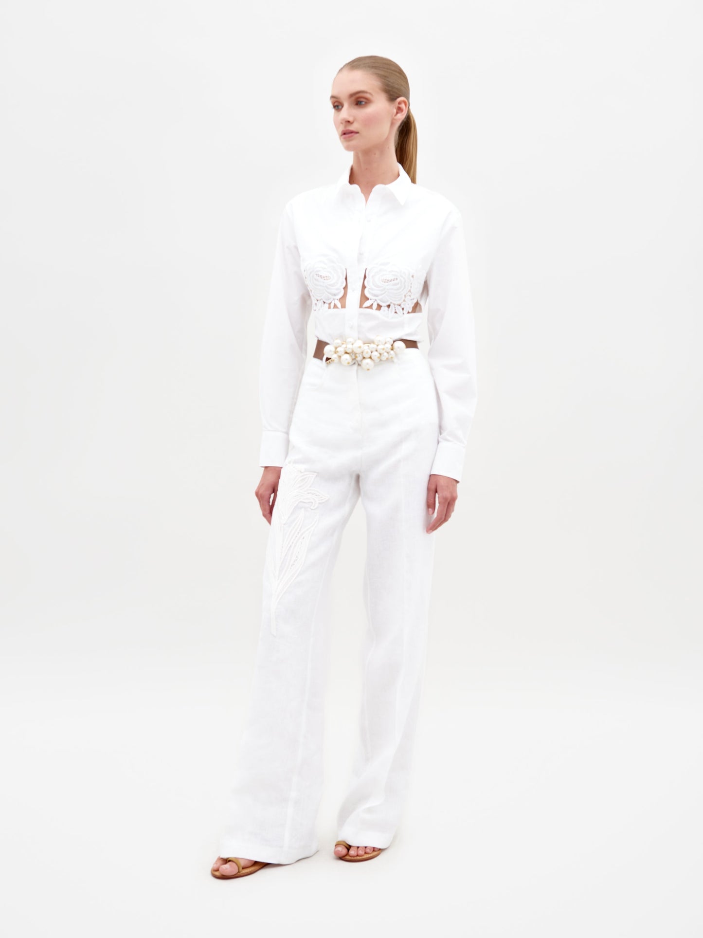 A person wearing the Sigrid Blouse White, a long-sleeved top featuring floral cut-outs, paired with high-waisted white pants with a floral belt, stands against a plain white background. Ready for pre-order, mark your calendars for February 15th, 2025, the anticipated ship-by date.