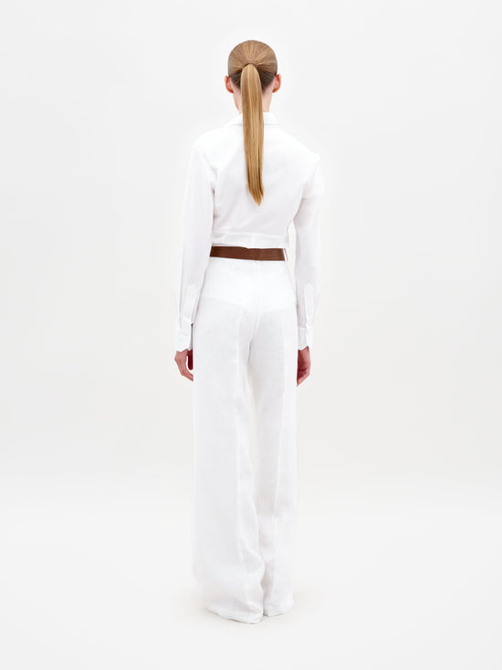 A person wearing the Sigrid Blouse White, a long-sleeved top featuring floral cut-outs, paired with high-waisted white pants with a floral belt, stands against a plain white background. Ready for pre-order, mark your calendars for February 15th, 2025, the anticipated ship-by date.