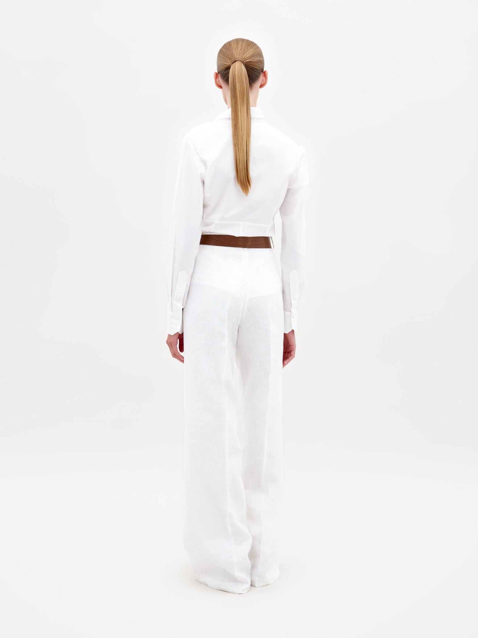 A person wearing the Sigrid Blouse White, a long-sleeved top featuring floral cut-outs, paired with high-waisted white pants with a floral belt, stands against a plain white background. Ready for pre-order, mark your calendars for February 15th, 2025, the anticipated ship-by date.