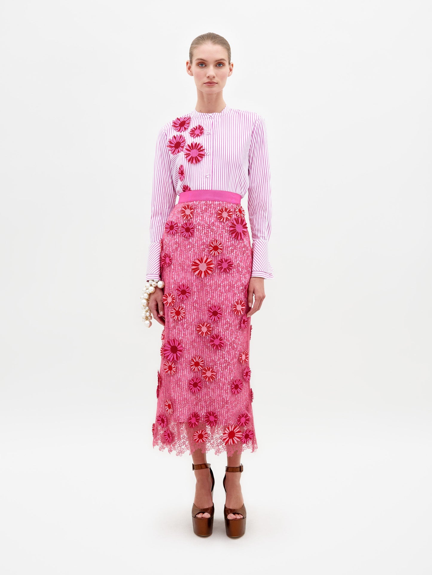 A woman stands wearing a long-sleeved, pink and white striped top paired with the floral-decorated, fuchsia Uma Skirt. She is also wearing brown high-heeled platform shoes. The background is plain white. Pre-order now to ensure your outfit ships by February 15th.