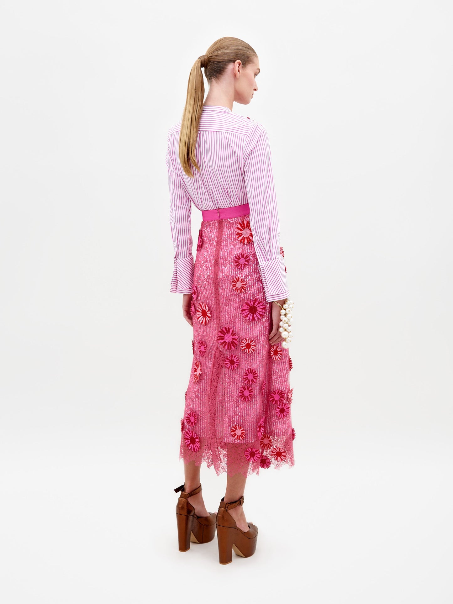 A woman stands wearing a long-sleeved, pink and white striped top paired with the floral-decorated, fuchsia Uma Skirt. She is also wearing brown high-heeled platform shoes. The background is plain white. Pre-order now to ensure your outfit ships by February 15th.