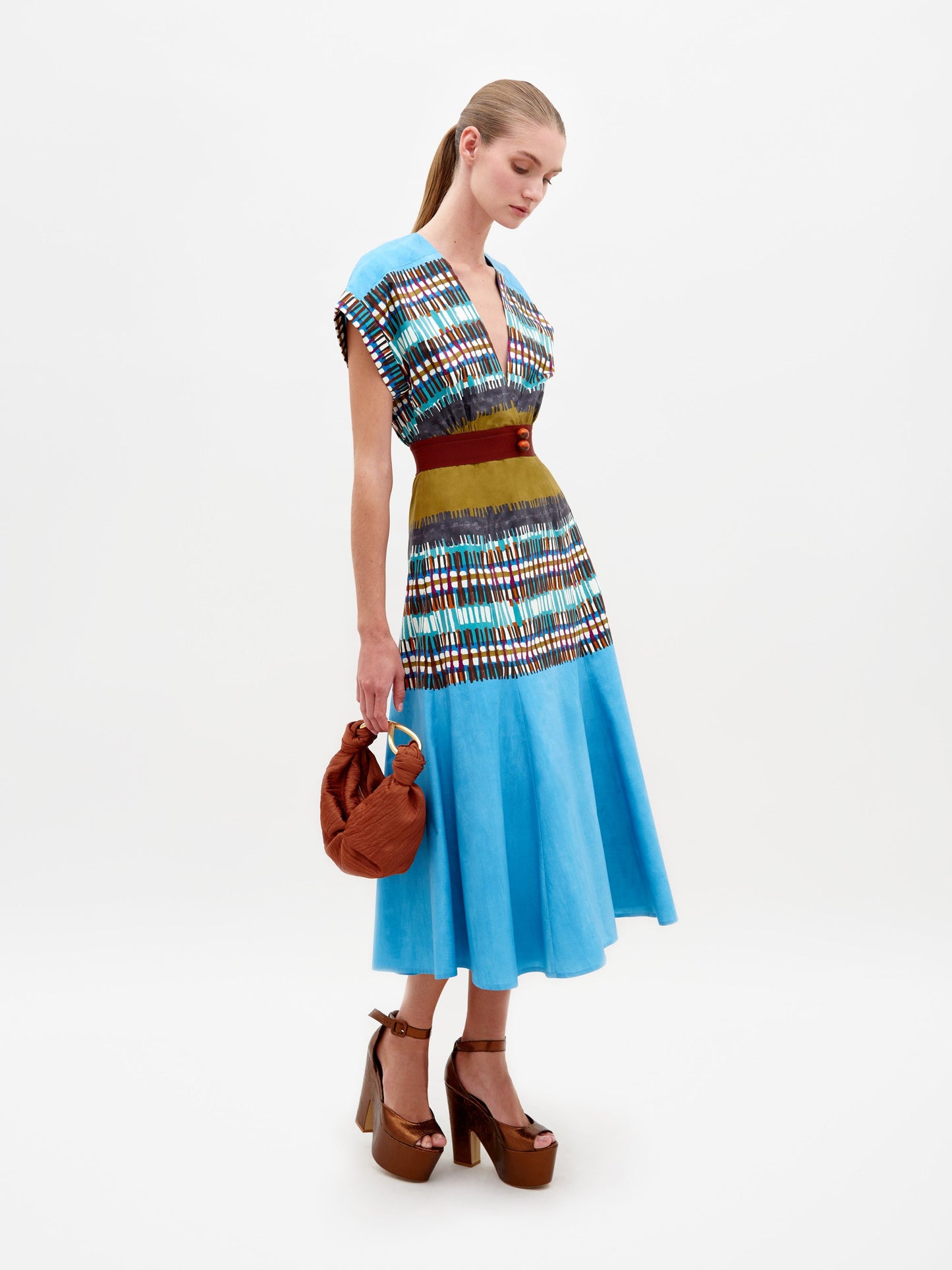 A woman stands wearing a Sakura Dress in Aqua Olive Plaid, featuring a V-neck design with multicolored patterns. She holds a brown handbag and wears brown platform heels. Available for pre-order, this look will ship by February 15th, 2025.