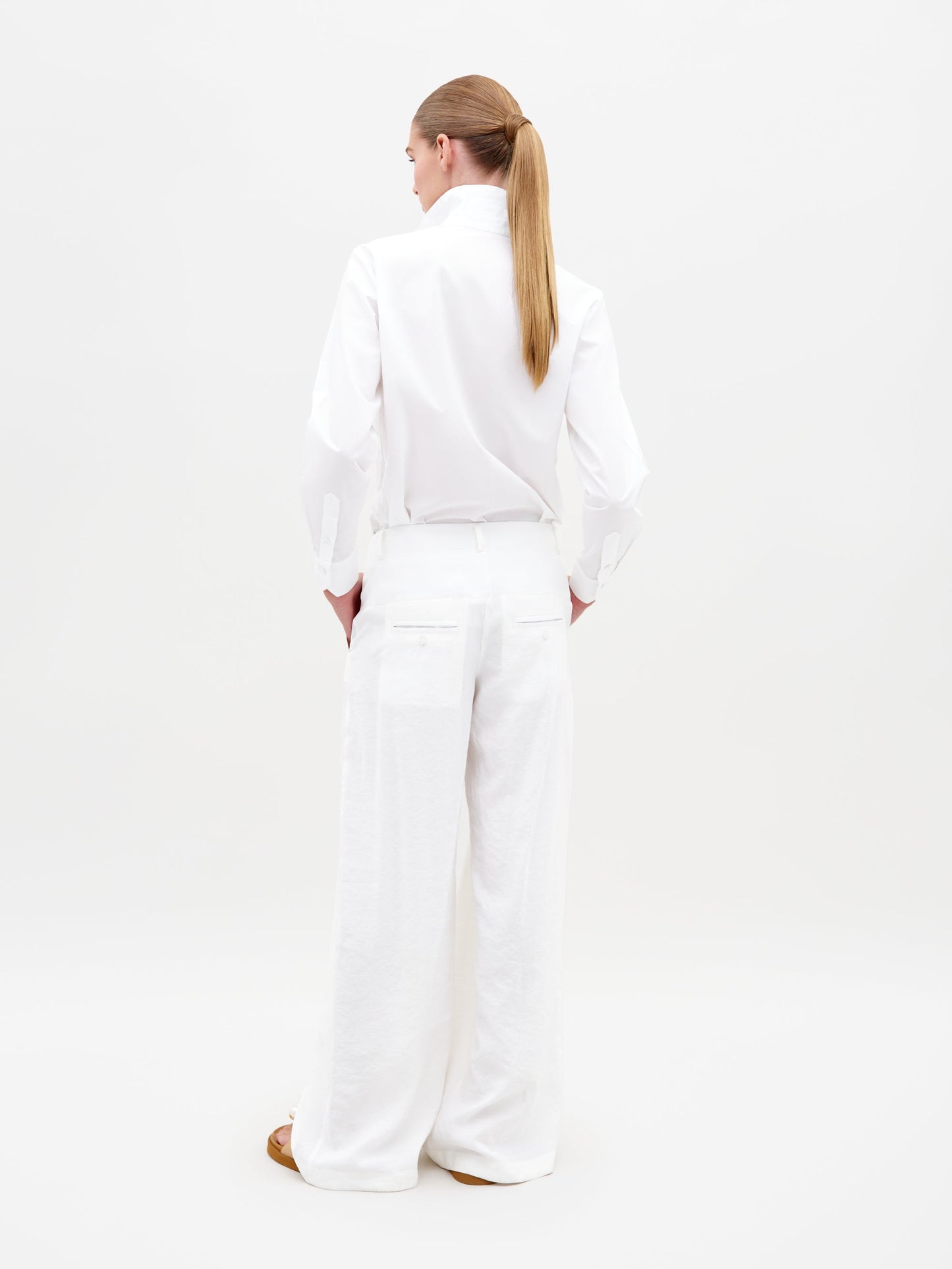 A person stands against a plain background, wearing an all-white outfit featuring the Opal Pant White—a pair of wide-legged pants—combined with a long-sleeve shirt with a high collar, their hands casually in their pockets. Pre-order now to secure your style for February 15th, 2025.