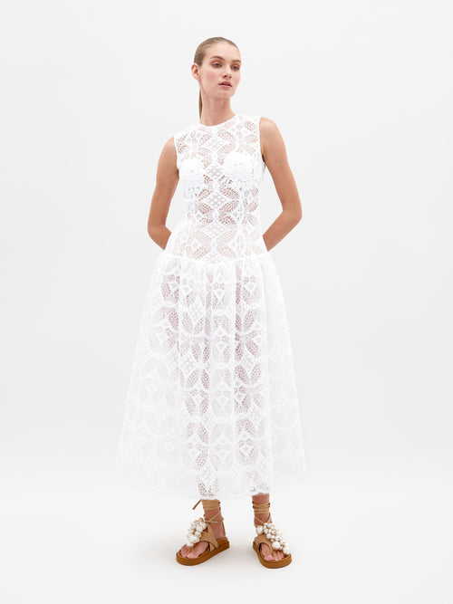 A person stands in the Paola Dress White, featuring a sleeveless lace design with a flared skirt, complemented by brown lace-up sandals. This item is available for pre-order and will ship by February 15th, 2025. The background is plain white.