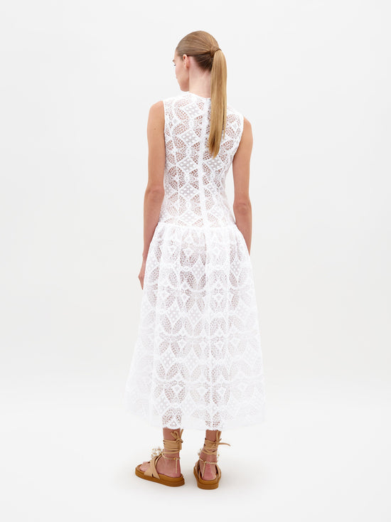 A person stands in the Paola Dress White, featuring a sleeveless lace design with a flared skirt, complemented by brown lace-up sandals. This item is available for pre-order and will ship by February 15th, 2025. The background is plain white.