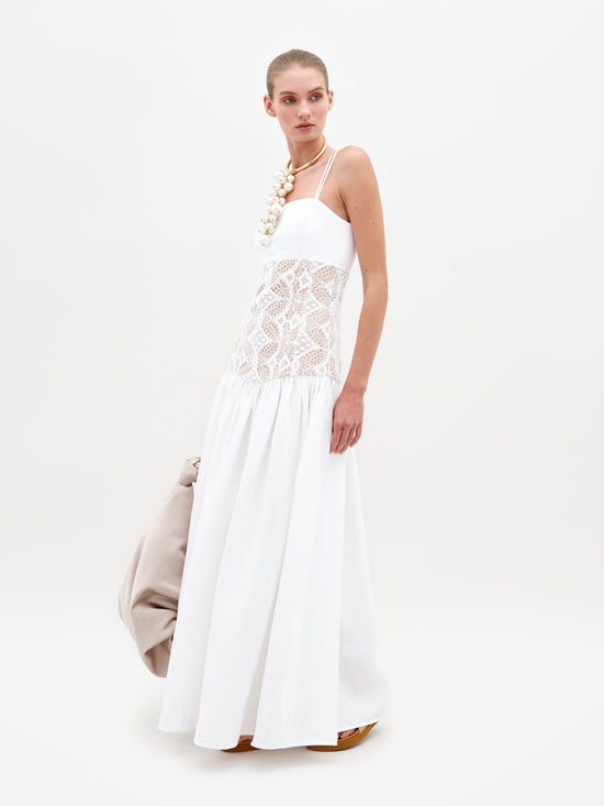 A woman stands wearing the Pilar Dress White, a sleeveless white dress featuring a lace midsection, and holds a beige bag against a plain white background. This elegant piece is available for pre-order and will be shipped by February 15th, 2025.