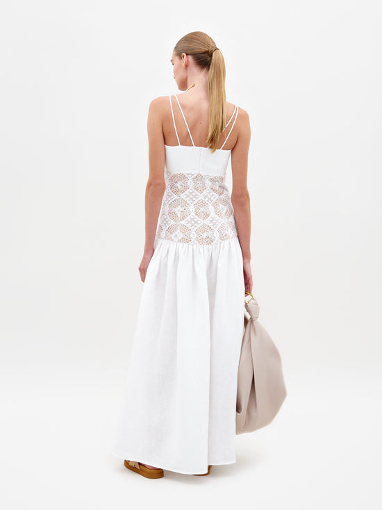 A woman stands wearing the Pilar Dress White, a sleeveless white dress featuring a lace midsection, and holds a beige bag against a plain white background. This elegant piece is available for pre-order and will be shipped by February 15th, 2025.