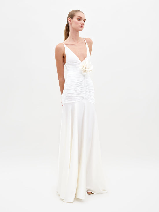 A woman in the Sana Dress White, featuring a sleeveless design with a deep V-neck and floral detail at the waist, stands against a plain white background. Pre-order now to ensure delivery by February 15th, 2025.