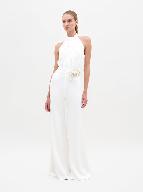 A woman stands against a plain background, wearing the Siv Jumpsuit White—a sleeveless halter jumpsuit featuring a beaded embellishment at the waist. This stunning piece is now available for pre-order and is set to ship by February 15th, 2025.