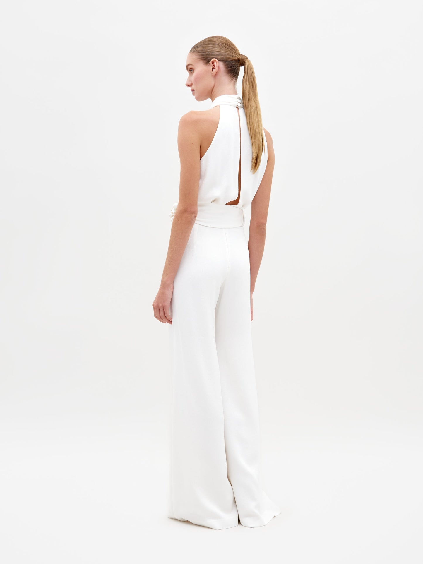 A woman stands against a plain background, wearing the Siv Jumpsuit White—a sleeveless halter jumpsuit featuring a beaded embellishment at the waist. This stunning piece is now available for pre-order and is set to ship by February 15th, 2025.