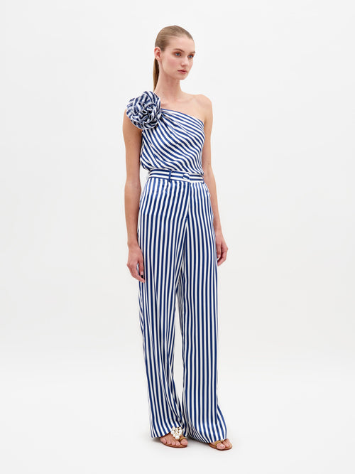 A woman stands wearing the Emine Pant White Blue Stripes, a one-shoulder jumpsuit featuring blue and white stripes and a large shoulder ruffle, available for pre-order until February 15th, 2025.