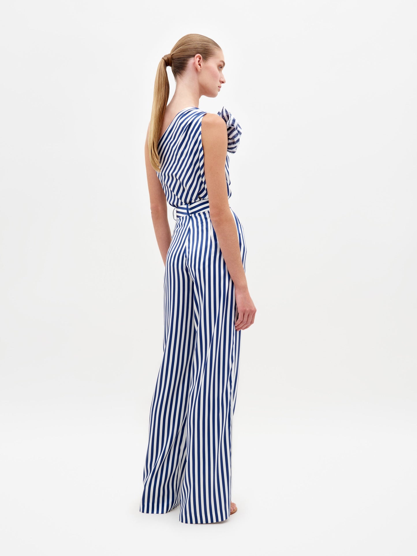 A woman stands wearing the Emine Pant White Blue Stripes, a one-shoulder jumpsuit featuring blue and white stripes and a large shoulder ruffle, available for pre-order until February 15th, 2025.