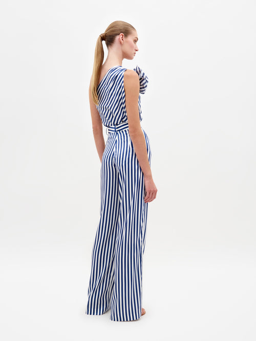 A woman stands wearing the Emine Pant White Blue Stripes, a one-shoulder jumpsuit featuring blue and white stripes and a large shoulder ruffle, available for pre-order until February 15th, 2025.