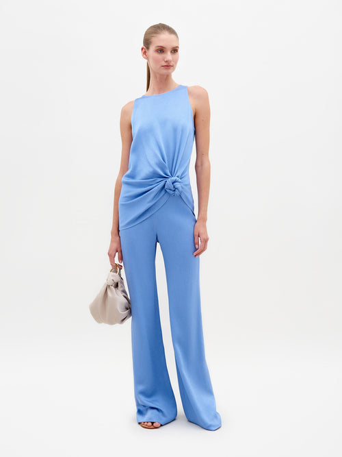 A woman wearing a sleeveless, periwinkle top with a knot at the waist and matching Palermo Pant in Periwinkle stands holding a beige handbag against a plain white background. Pre-order now for delivery by February 15th, 2025!