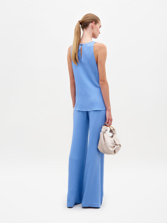 A woman wearing a sleeveless, periwinkle top with a knot at the waist and matching Palermo Pant in Periwinkle stands holding a beige handbag against a plain white background. Pre-order now for delivery by February 15th, 2025!