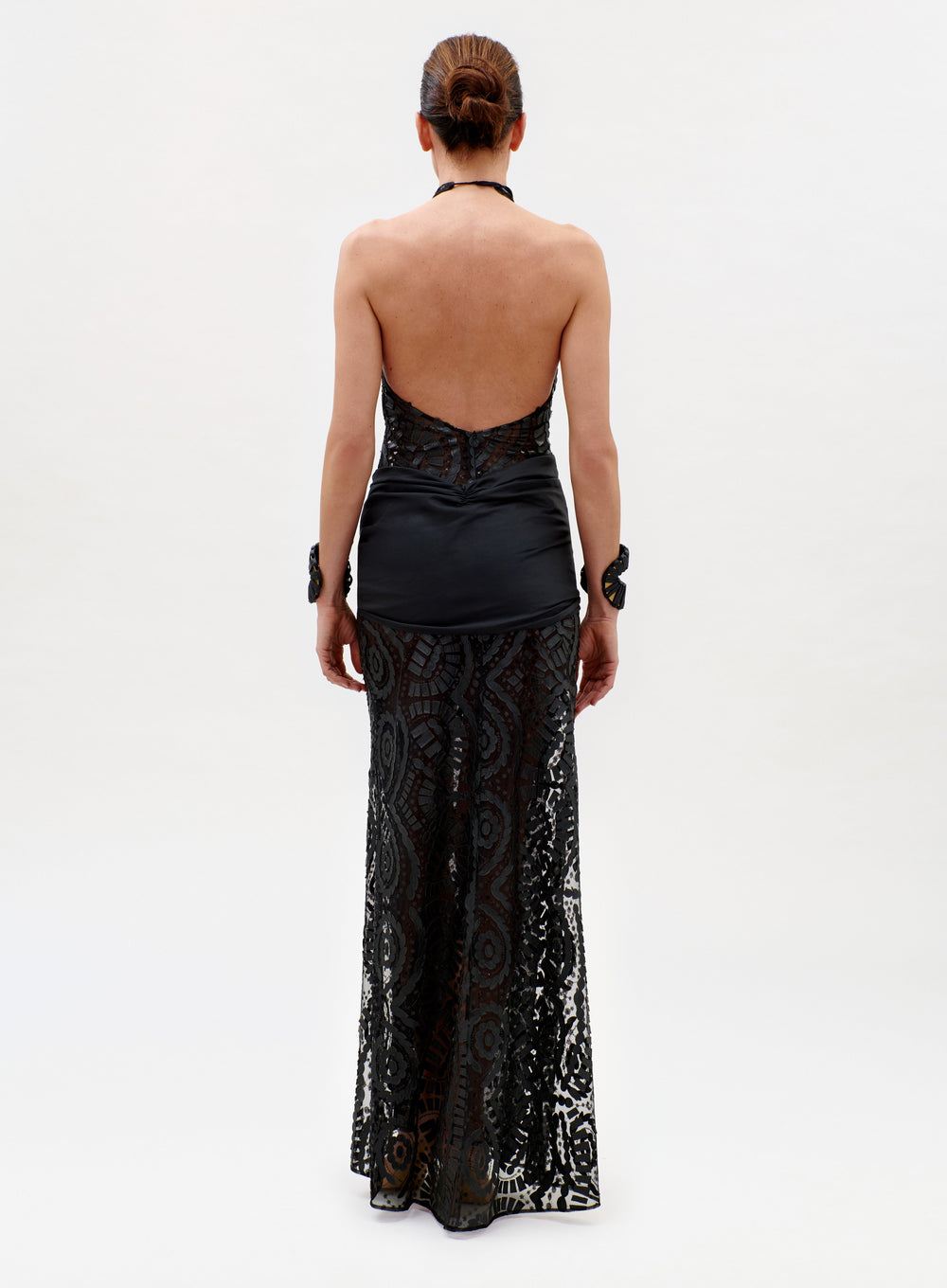 A person, facing away, wears the Lili Dress—a black halter-neck style with a sheer lace skirt, satin fitted top, open back, and two matching lace cuffs—against a plain white background.