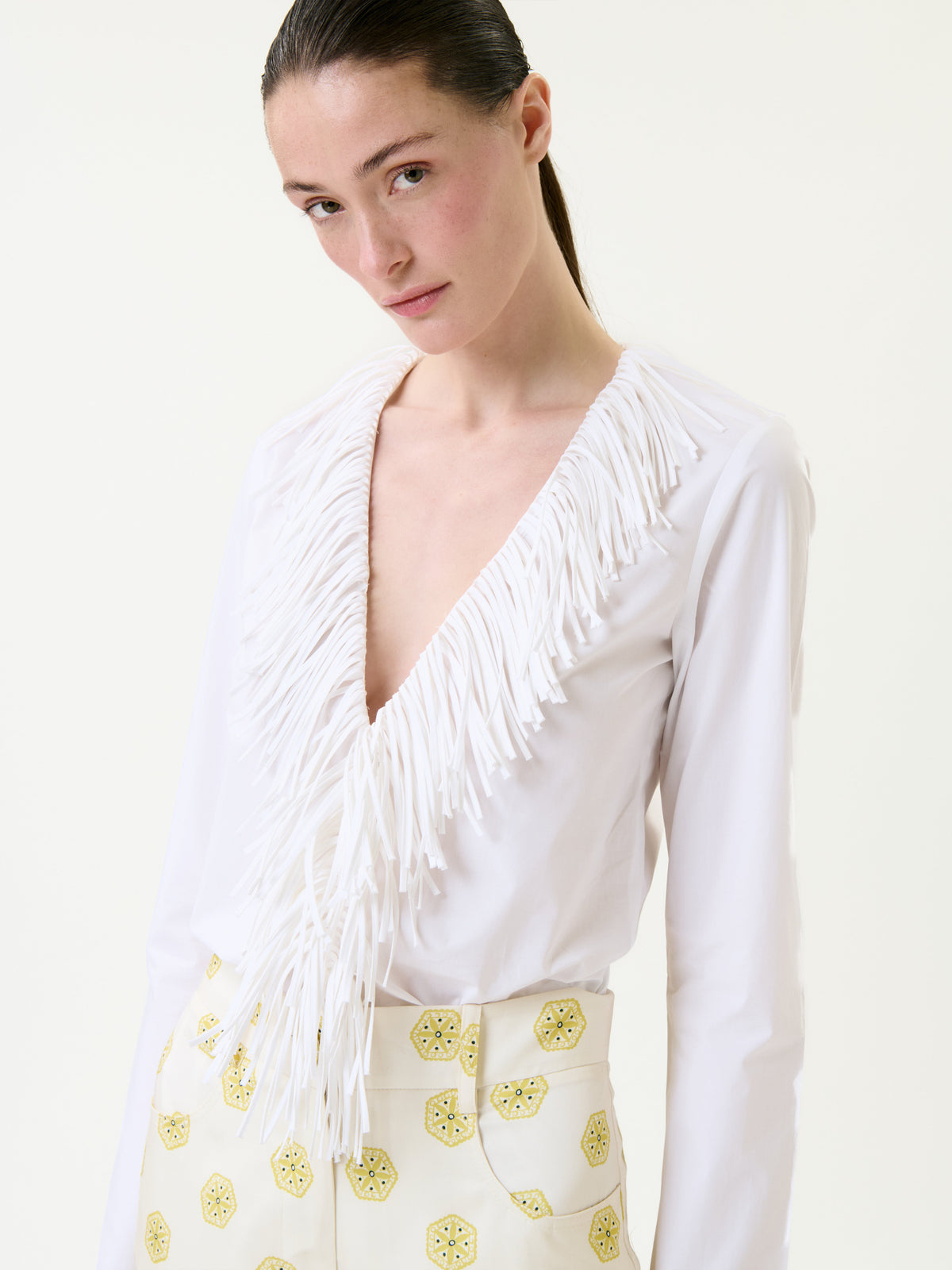 A person wearing the Zena Shirt, a white long-sleeve deep V-neck top with fringe details, and high-waisted cream pants with yellow patterns stands against a plain light background.