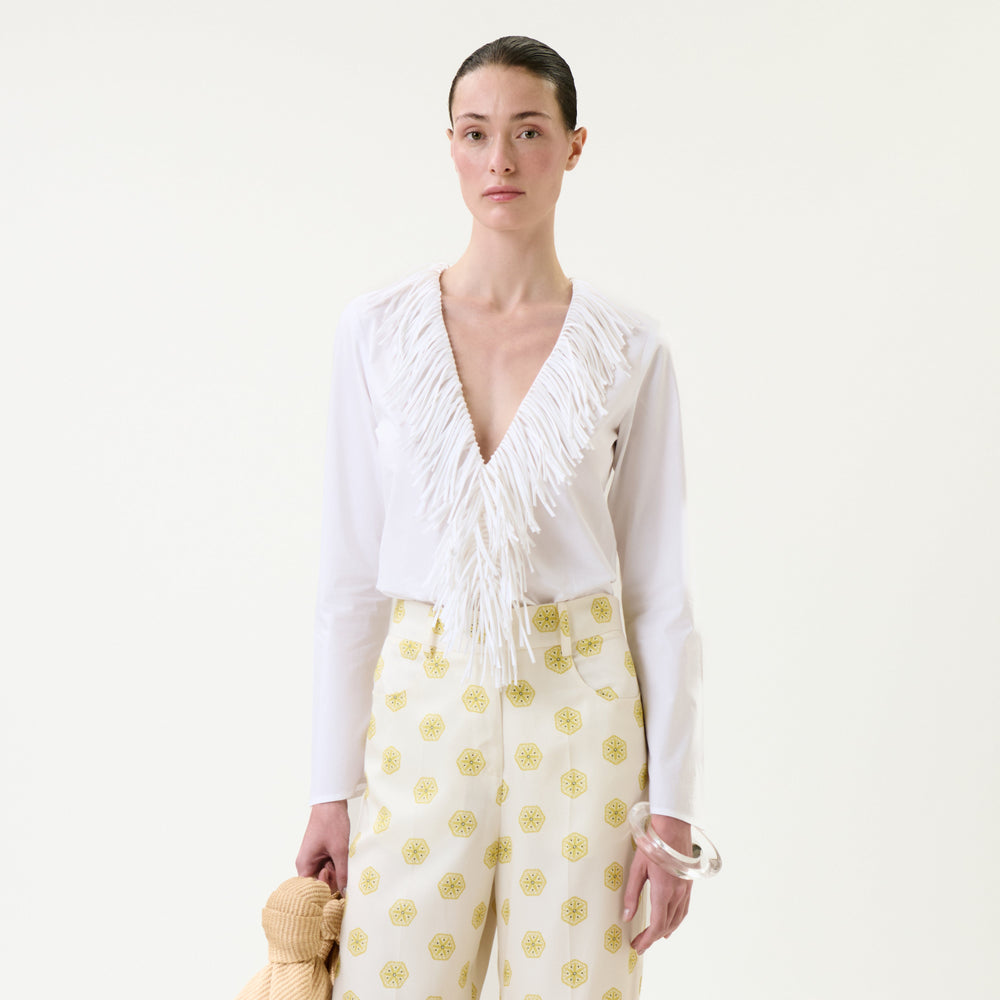 A woman stands against a plain background, wearing the Zena Shirt—a white V-neck blouse with fringe—paired with high-waisted white pants featuring yellow patterns; she holds a textured beige item and accessorizes with a silver bangle.