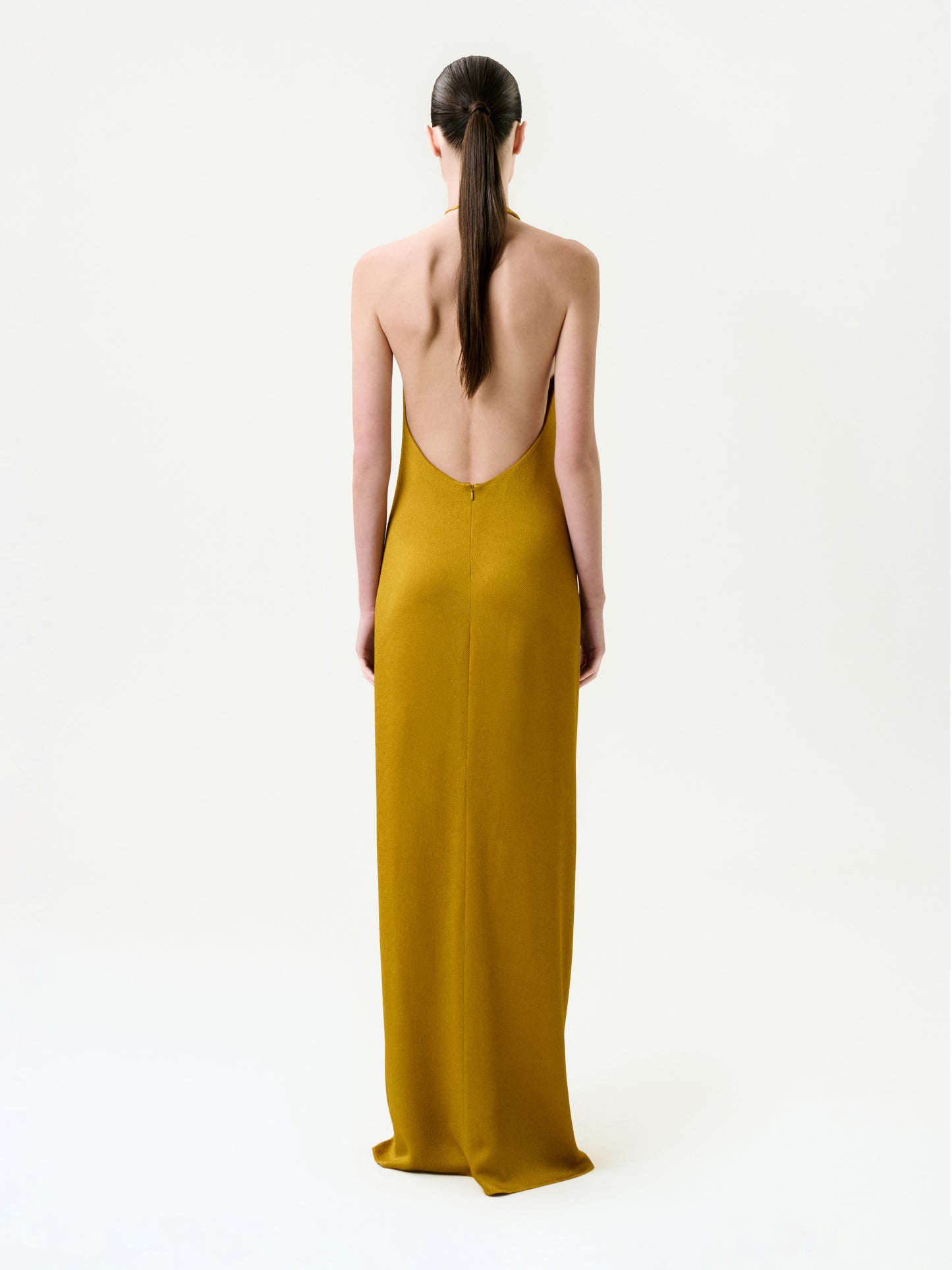 A person with long, straight dark hair in a low ponytail wears the Sira Dress—a backless, floor-length mustard yellow maxi crafted from Italian fabric—standing against a plain white background, facing away from the camera.