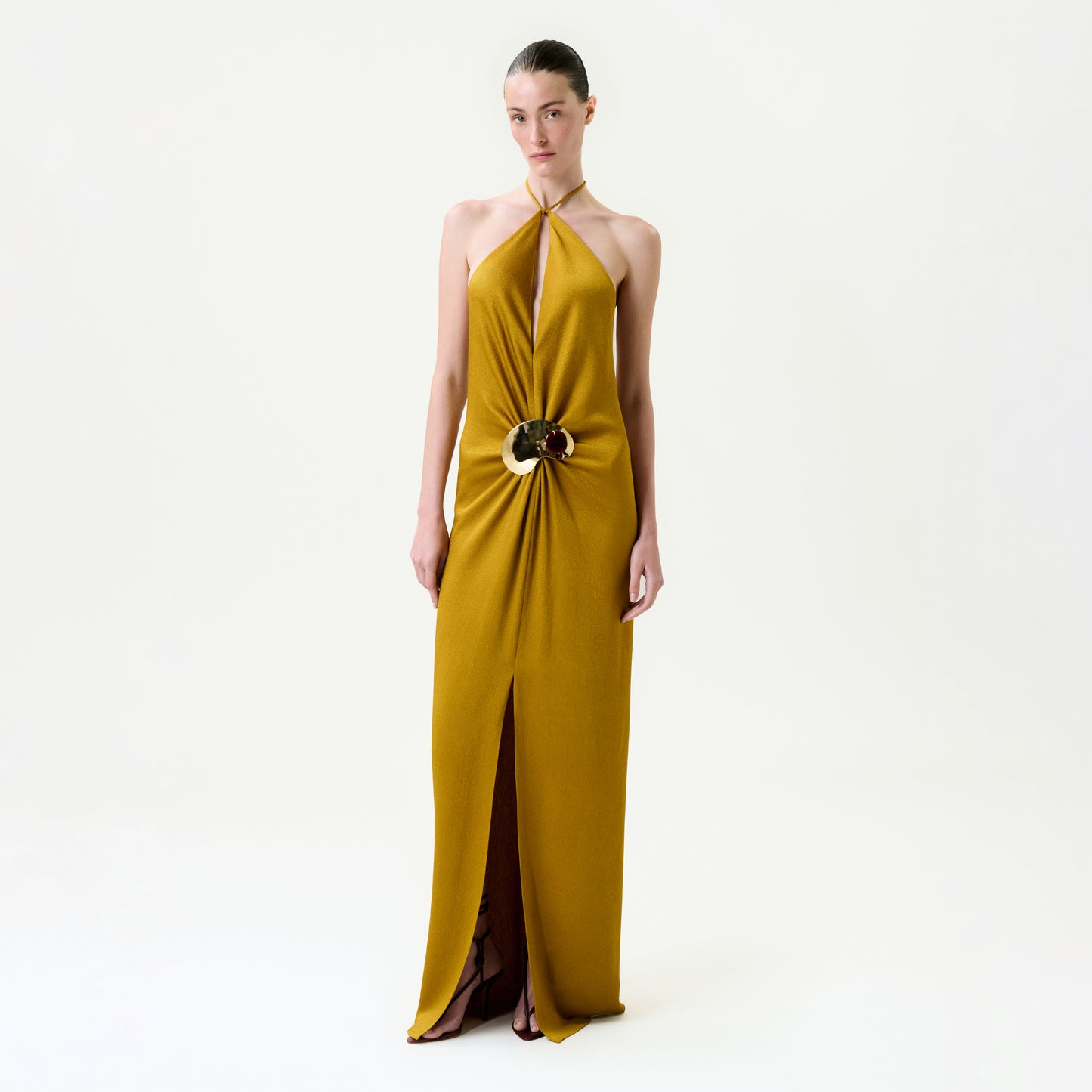 A woman stands against a plain white background wearing the Sira Dress—a mustard yellow maxi made from luxurious Italian fabric, featuring a halter-neck, keyhole neckline, central gold and brown embellishment, and front slit.