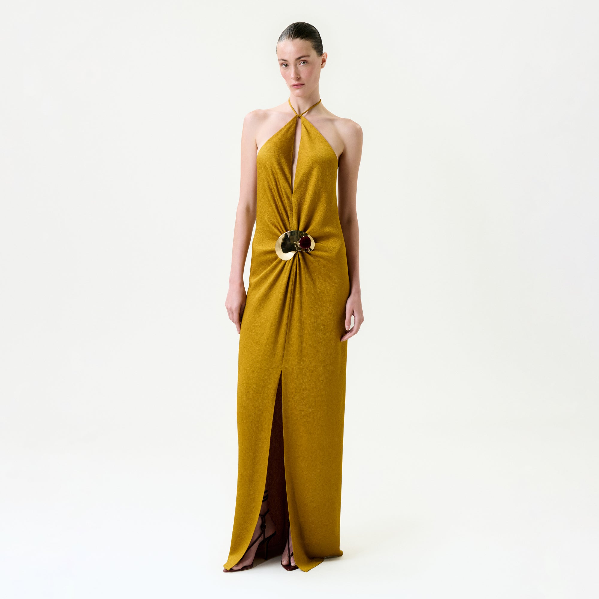 A woman stands against a plain white background wearing the Sira Dress—a mustard yellow maxi made from luxurious Italian fabric, featuring a halter-neck, keyhole neckline, central gold and brown embellishment, and front slit.