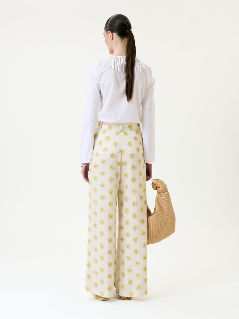 A person stands facing away, wearing a white blouse paired with the Emine Pant—wide-leg white pants featuring a yellow circular pattern—and holding a tan woven bag.