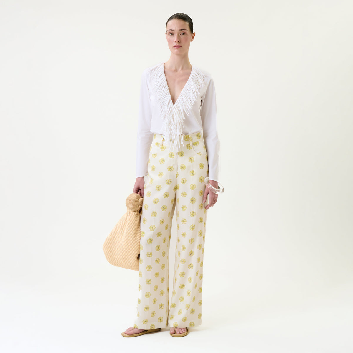 A woman stands against a plain background wearing a white blouse with a deep V and fringe detail, the Emine Pant – high-waisted wide-leg trousers featuring a yellow dot pattern, pink sandals, and holding a large beige textured bag.