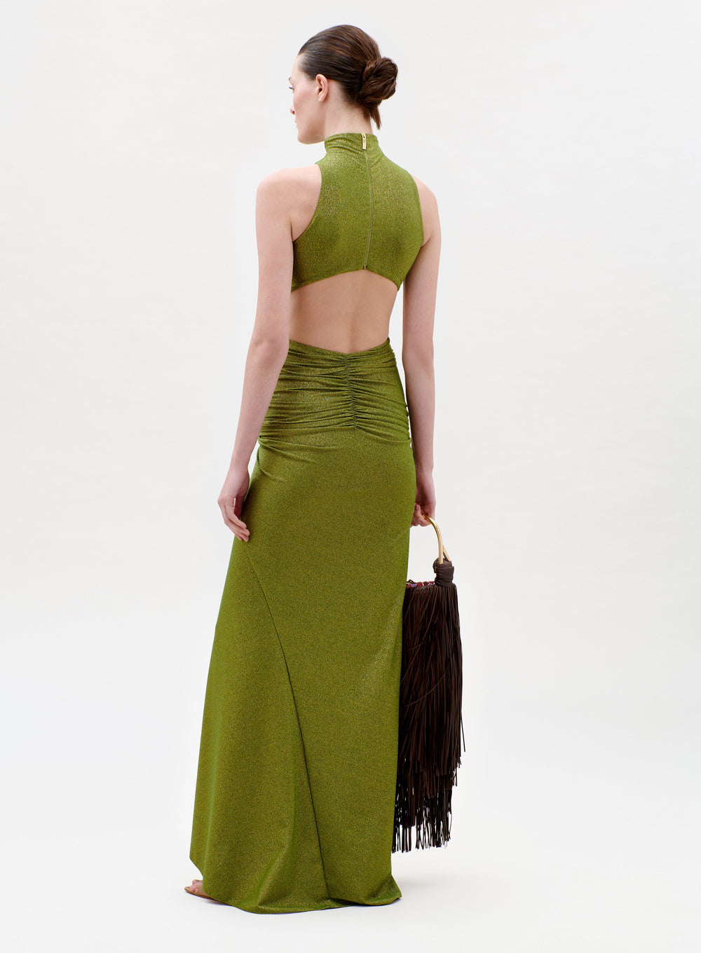 A woman in the Frances Dress—an olive green, sleeveless gown with an open back and ruched waist—stands barefoot holding a large brown fringed bag. Her hair is styled in a low bun against a plain light background.