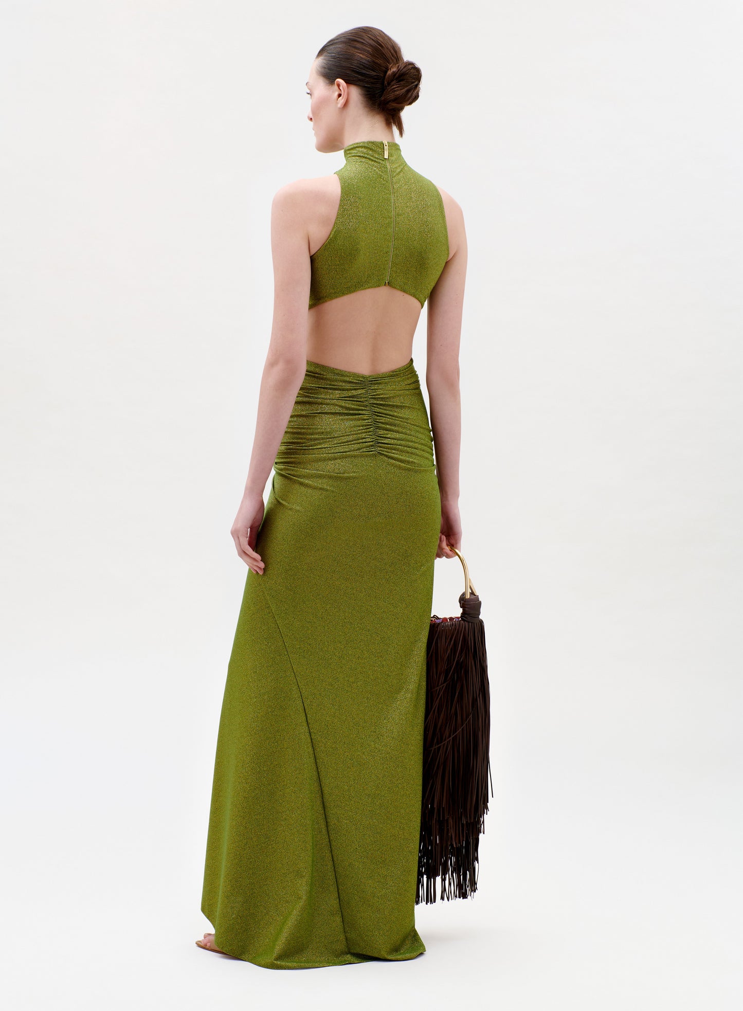 A woman in the Frances Dress—an olive green, sleeveless gown with an open back and ruched waist—stands barefoot holding a large brown fringed bag. Her hair is styled in a low bun against a plain light background.