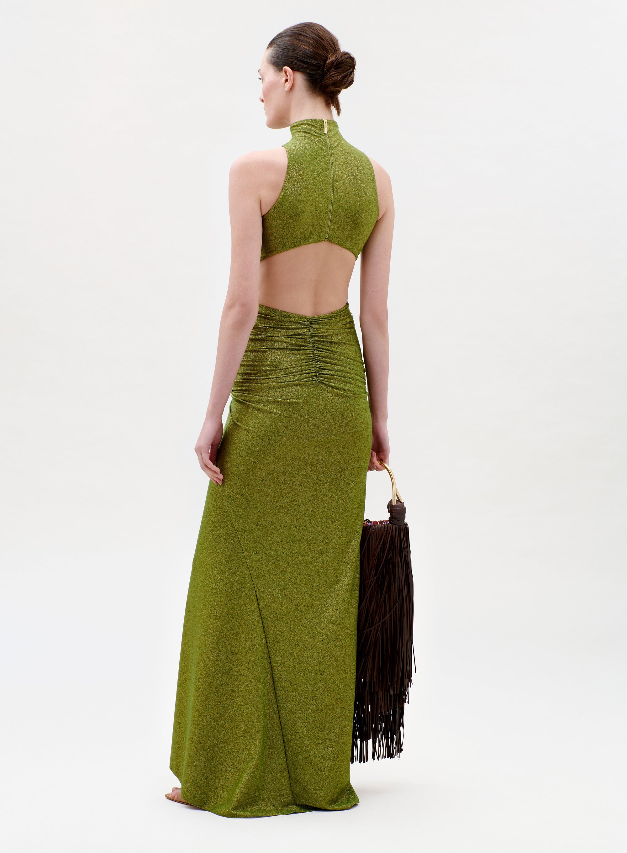 A woman in the Frances Dress—an olive green, sleeveless gown with an open back and ruched waist—stands barefoot holding a large brown fringed bag. Her hair is styled in a low bun against a plain light background.