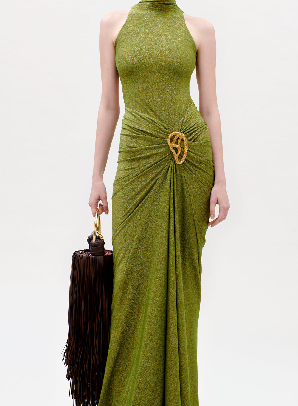 A person wearing the Frances Dress—a sleeveless, shimmering green gown with a high neckline and gold accent at the hip—holds a brown fringed handbag against a plain white background.