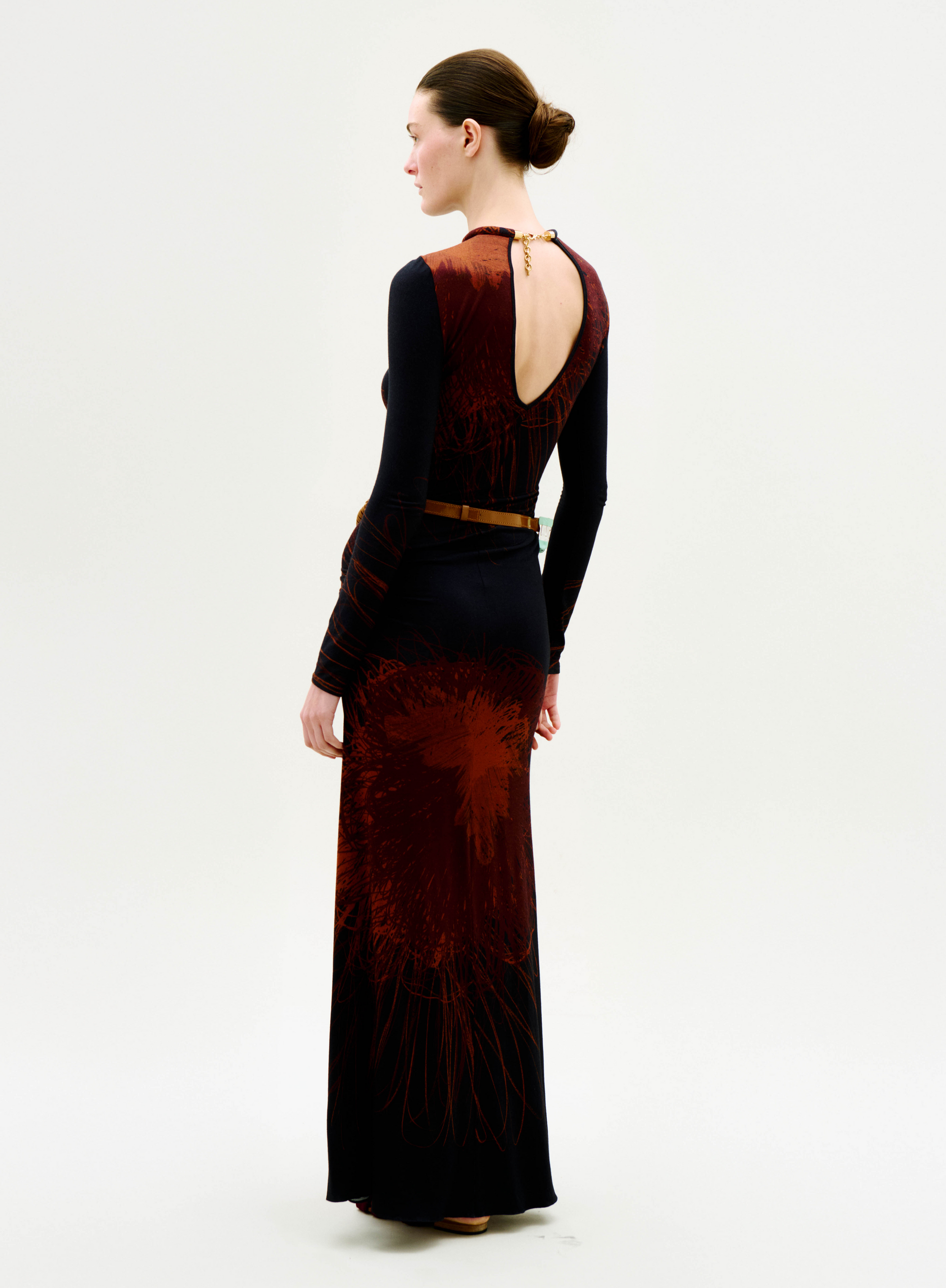 A woman with her hair in a neat bun stands against a plain white background, wearing the Gabbice Dress—a long-sleeved black and red patterned dress featuring an open back and a belt.