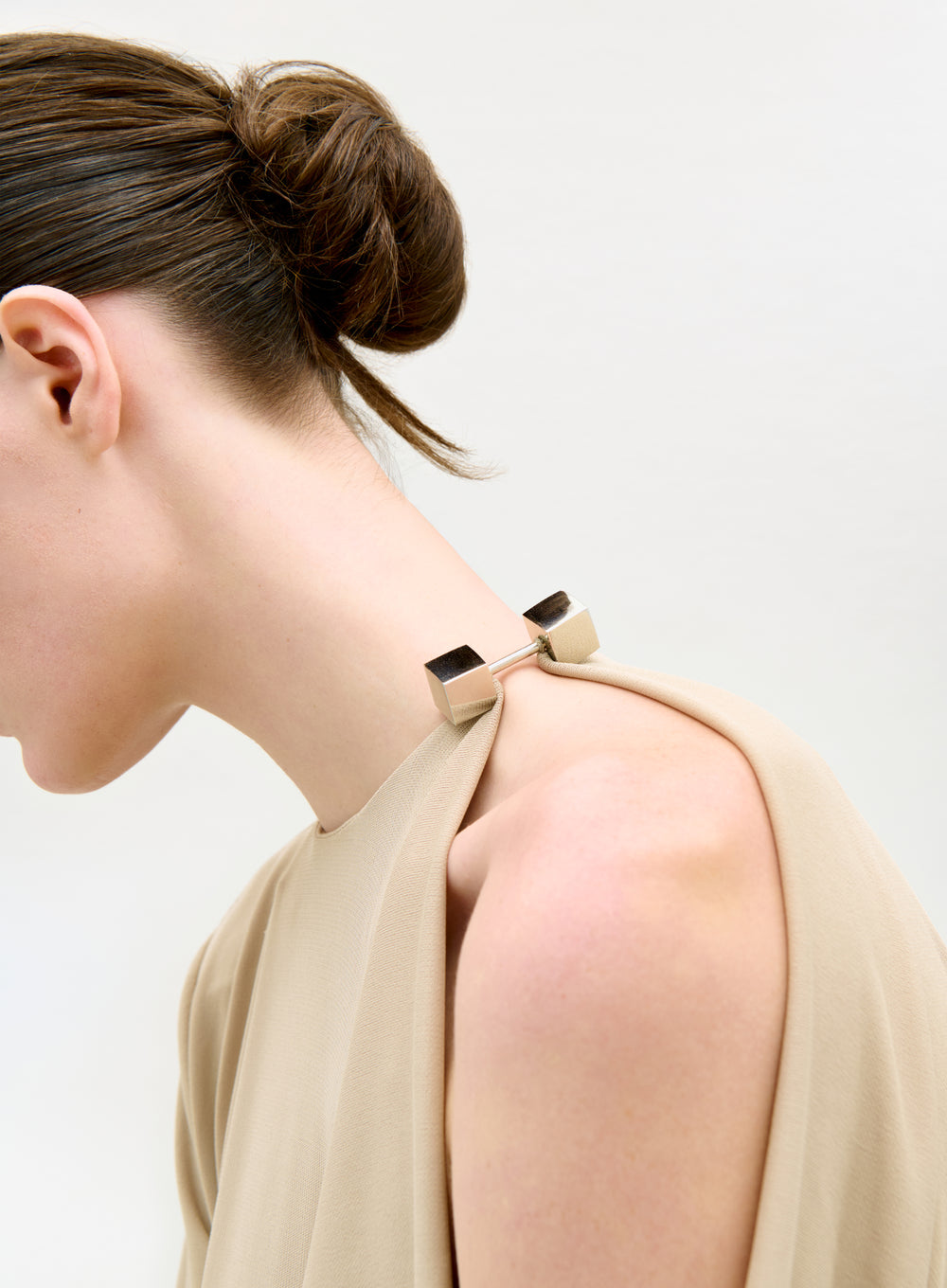 A person with dark hair in a sleek bun models the Natalia Dress—a beige, sleeveless piece fastened at the shoulder with a silver rectangular clasp—against a plain, light background.