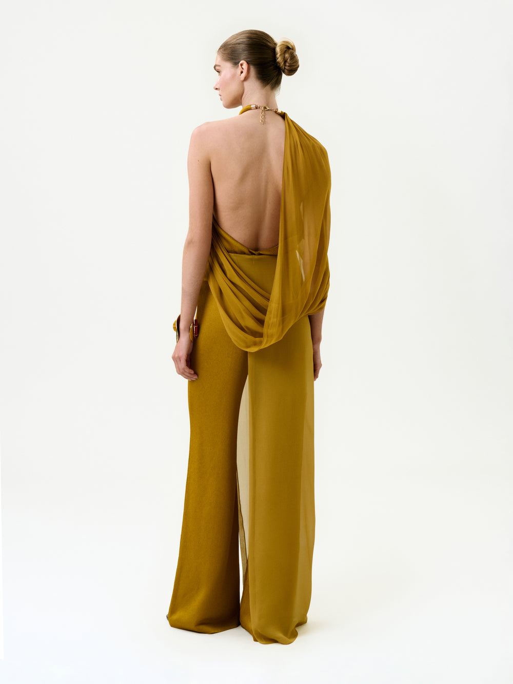 A woman stands facing away in the Gaucin Pant—a mustard yellow, backless halter-neck jumpsuit made from luxurious Italian fabric with a draped, silk-like texture and wide legs. Her hair is styled in a neat bun against a plain white background.