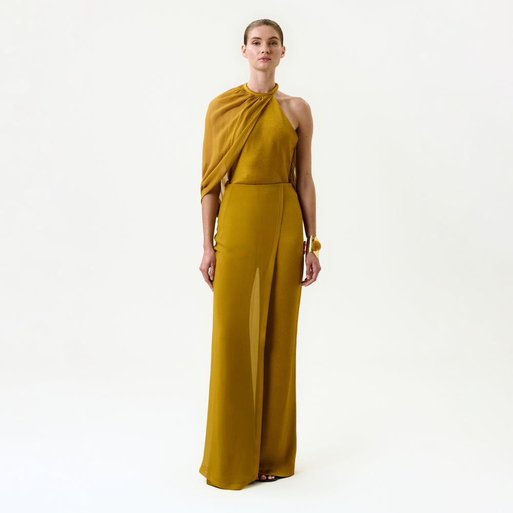 A woman stands against a plain white background wearing the Gaucin Pant in mustard yellow, a wide-leg style crafted from luxurious Italian fabric. Her hair is pulled back, and she maintains a neutral expression.
