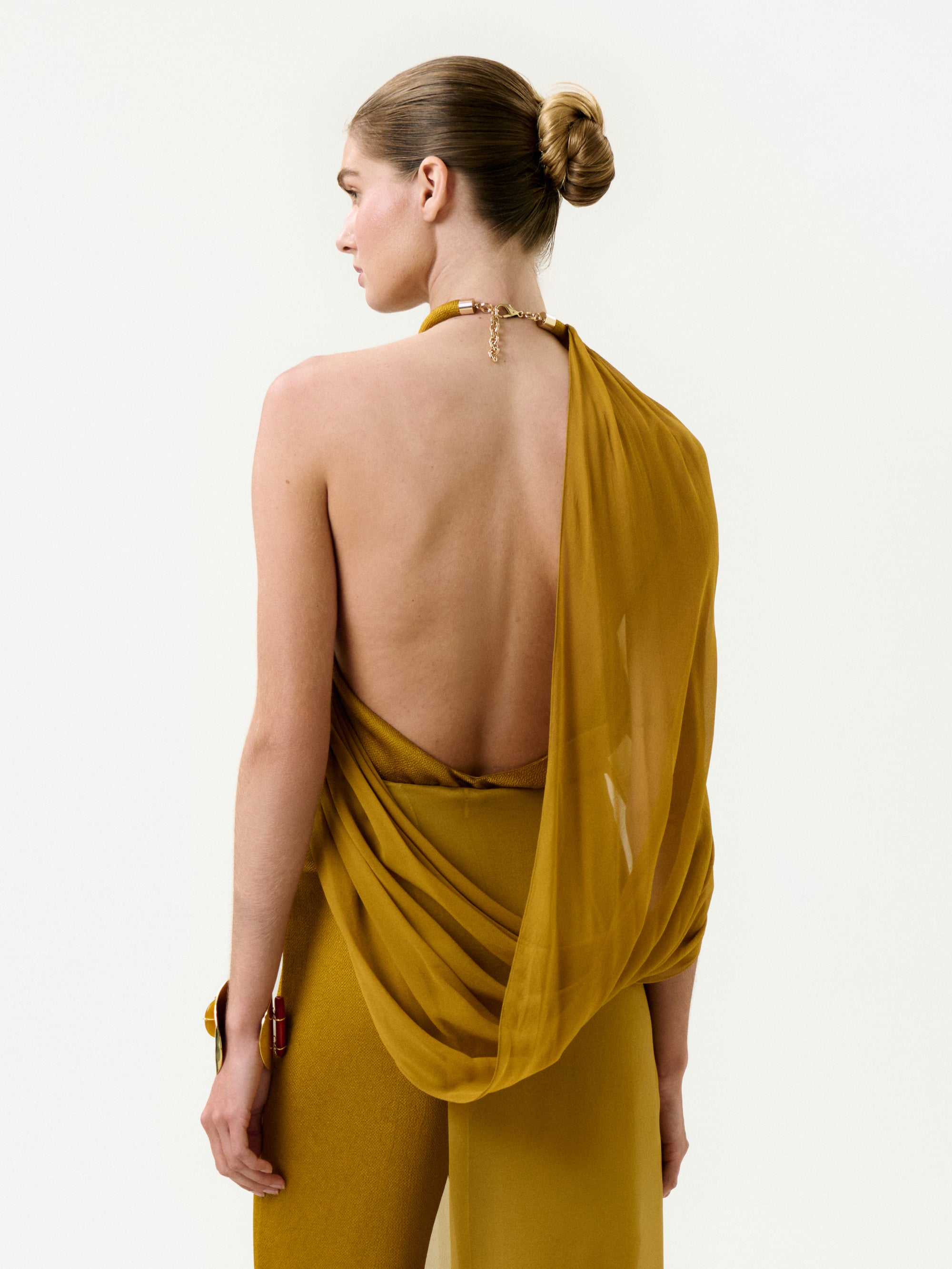 A woman stands facing away, wearing mustard yellow pants and the Milka Blouse—a draped, backless silk top. Her hair is in a sleek bun, and the luxurious fabric of the blouse hangs elegantly over one shoulder.