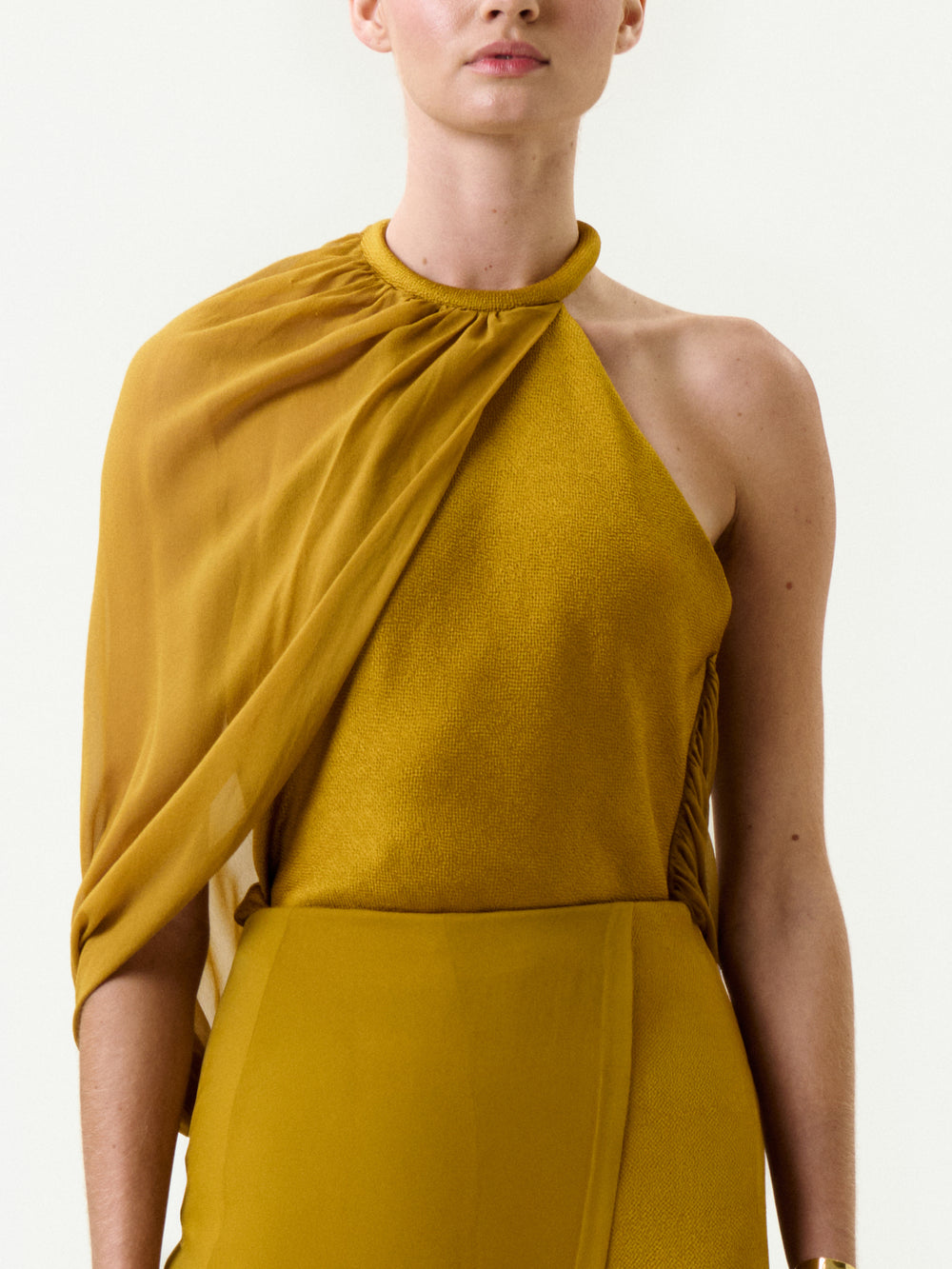 A person wearing the Milka Blouse—a mustard yellow, asymmetrical Italian fabric halter top with a sheer draped sleeve—paired with matching high-waisted pants, stands against a plain white background.