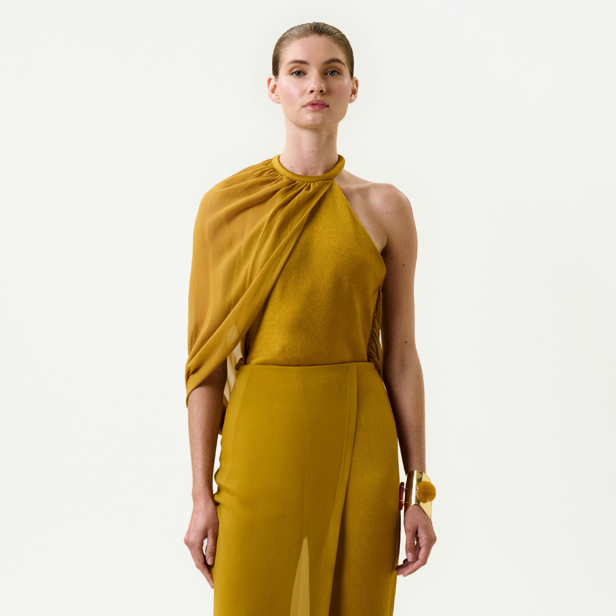 A woman stands against a white background, confidently wearing the Milka Blouse in mustard yellow Italian fabric. She holds a matching clutch, sports a gold bracelet, and her slicked-back hair completes the elegant look.