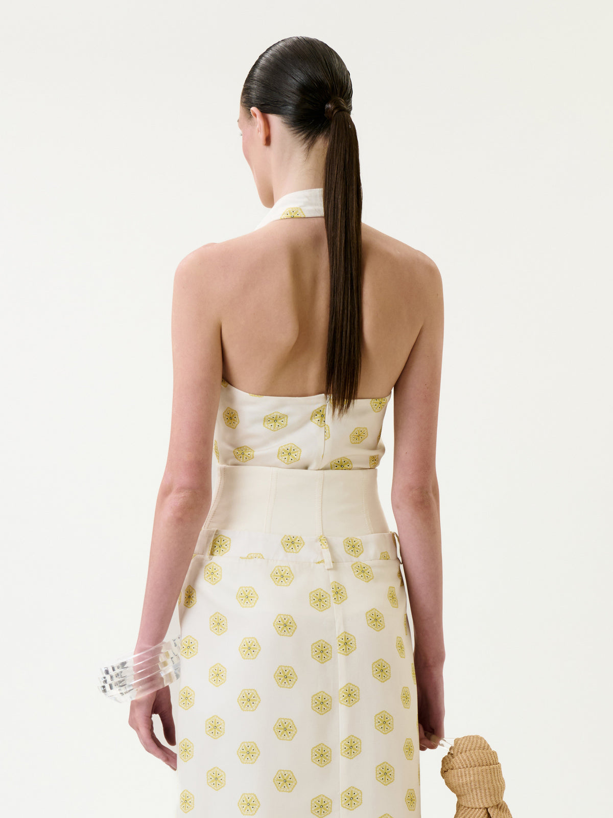 A woman with straight, dark hair in a ponytail is seen from the back wearing the Marine Blouse, a white top adorned with yellow circular patterns. She carries a tan woven bag and has a clear bracelet on her left wrist.