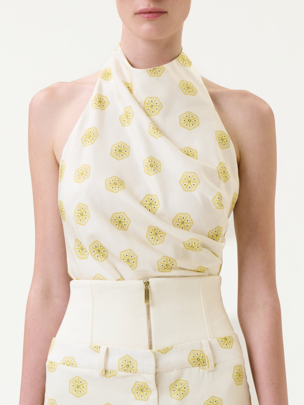 A woman wearing the Marine Blouse—a sleeveless cream halter top—paired with matching high-waisted pants featuring a yellow geometric pattern and a front zipper.