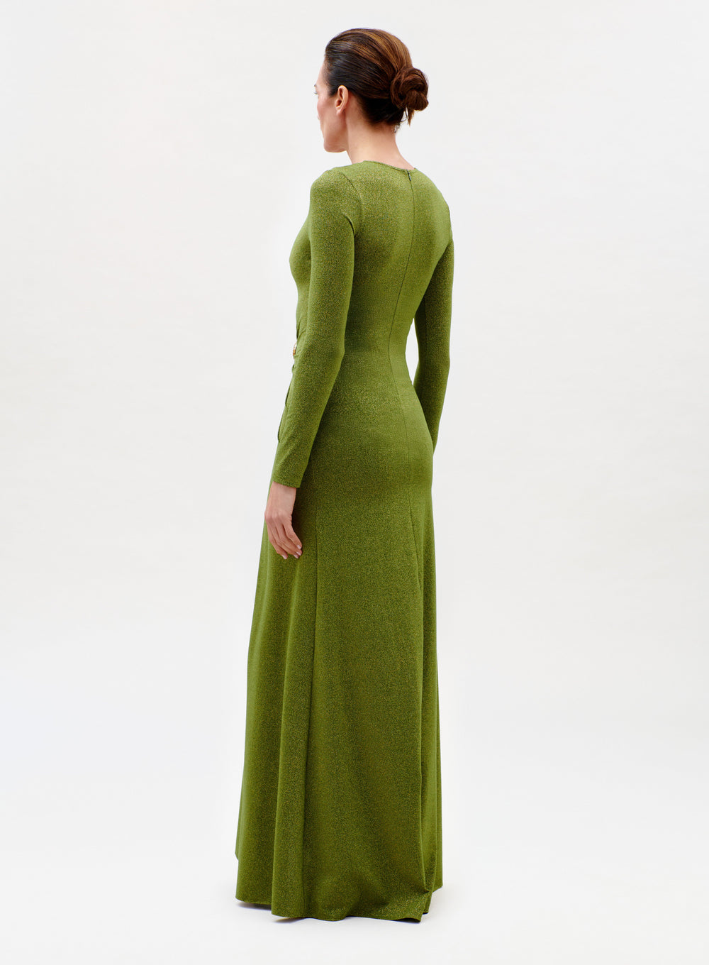 A woman faces away, wearing the Melany Dress—a long-sleeve, floor-length green design—with her hair in a neat bun against a plain light background.