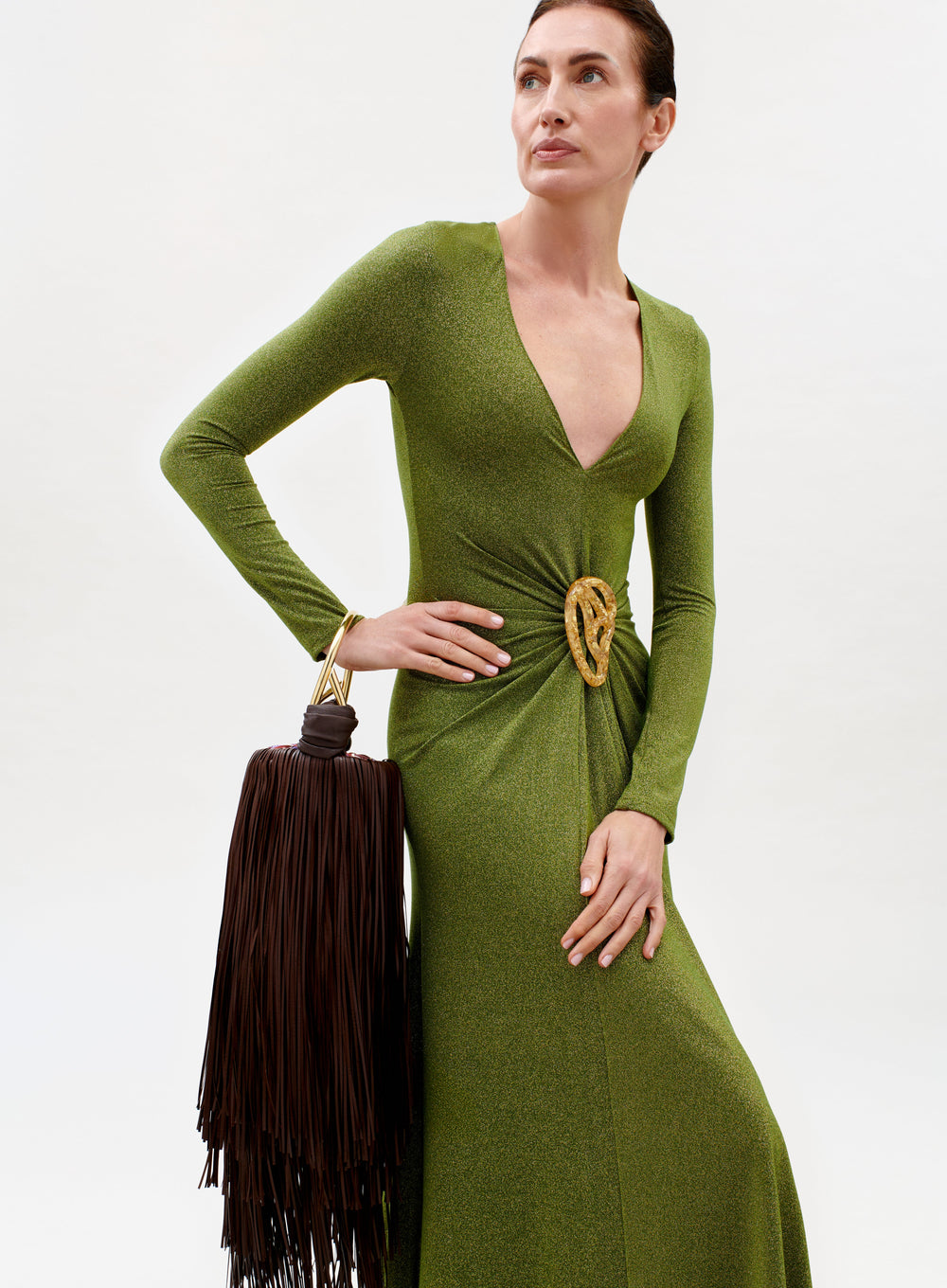 A woman in the Melany Dress—shimmering green with long sleeves and a deep V-neckline—poses against a plain background, holding a large brown fringed handbag and wearing a gold belt with a circular buckle.