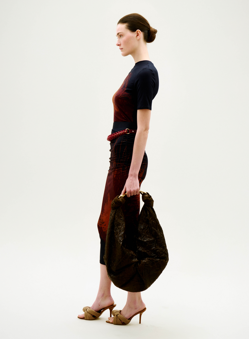 A woman in profile stands against a white background, wearing the Fano T-Shirt, heeled sandals, and holding a large crinkled brown bag. Her hair is pulled back in a neat bun.