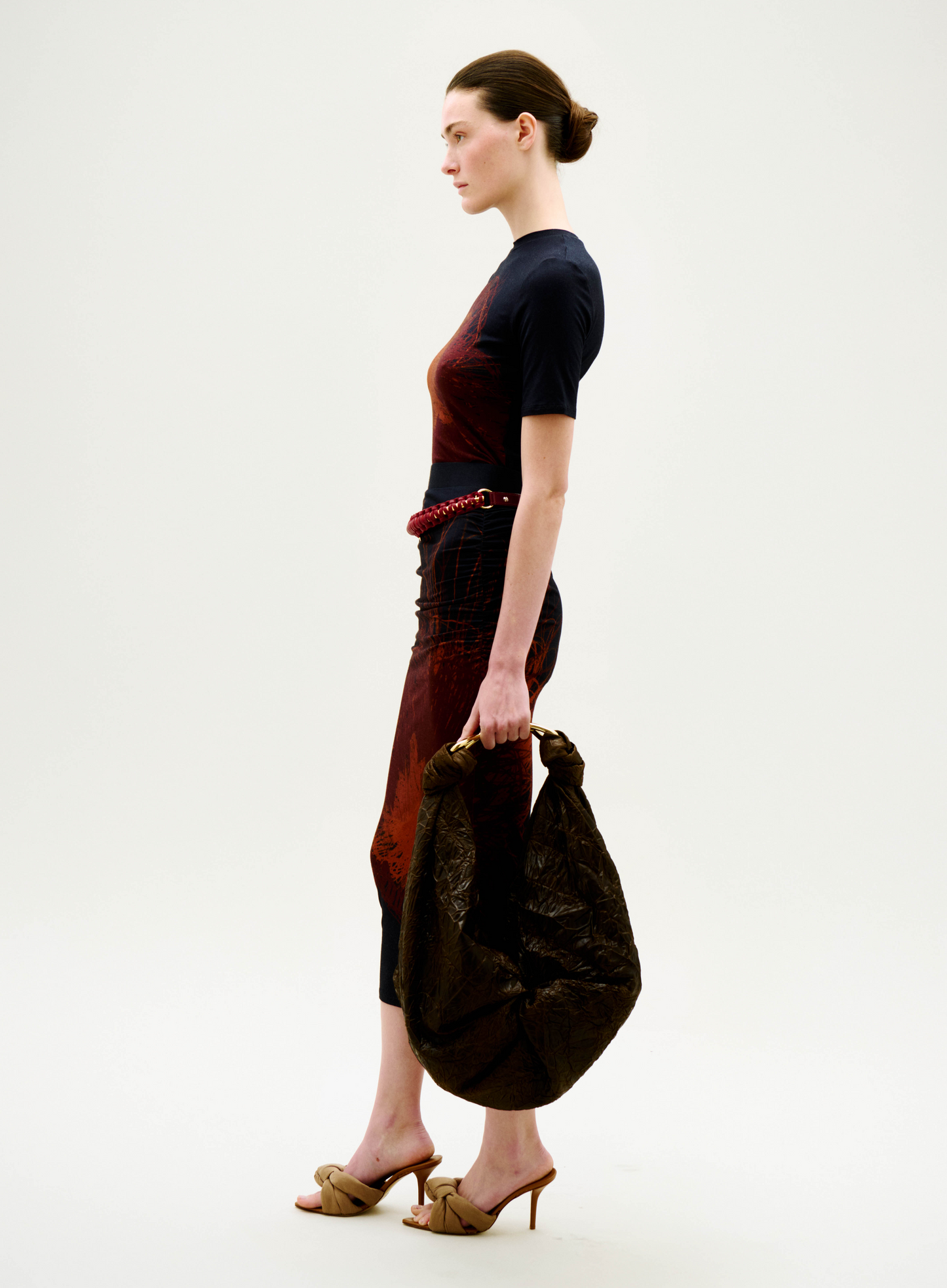 A woman in profile stands against a white background, wearing the Fano T-Shirt, heeled sandals, and holding a large crinkled brown bag. Her hair is pulled back in a neat bun.