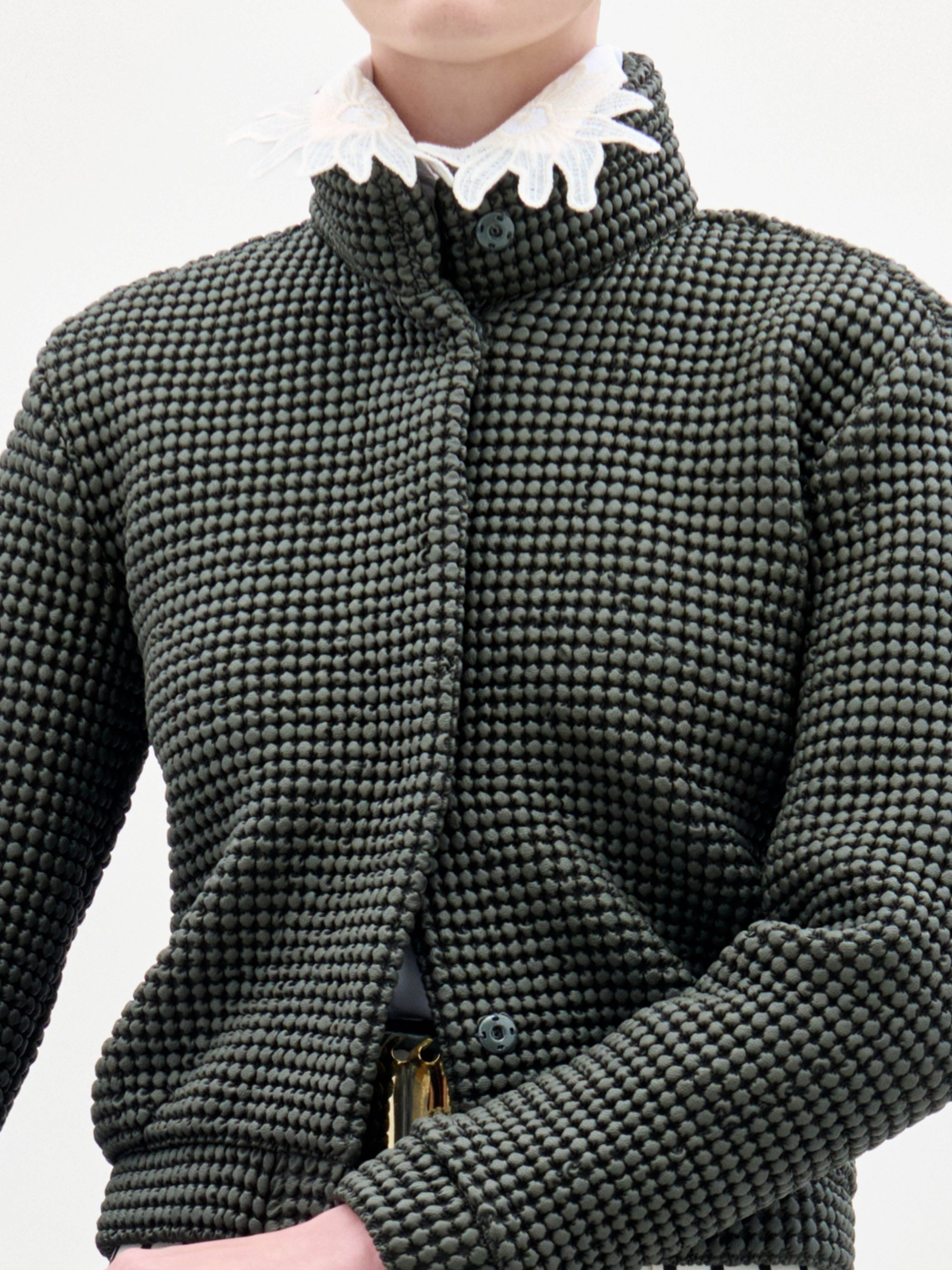 A woman with a neutral expression stands against a plain background wearing the textured Zalta Jacket, patterned pants, and holding a dark bag.