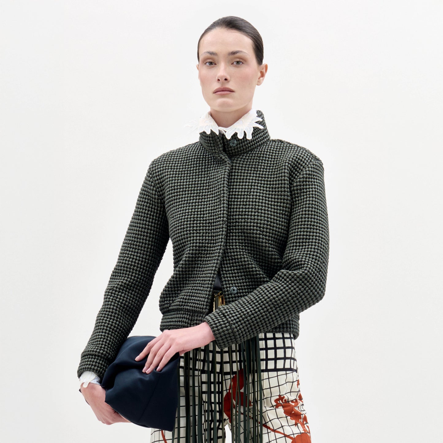 A woman with a neutral expression stands against a plain background wearing the textured Zalta Jacket, patterned pants, and holding a dark bag.