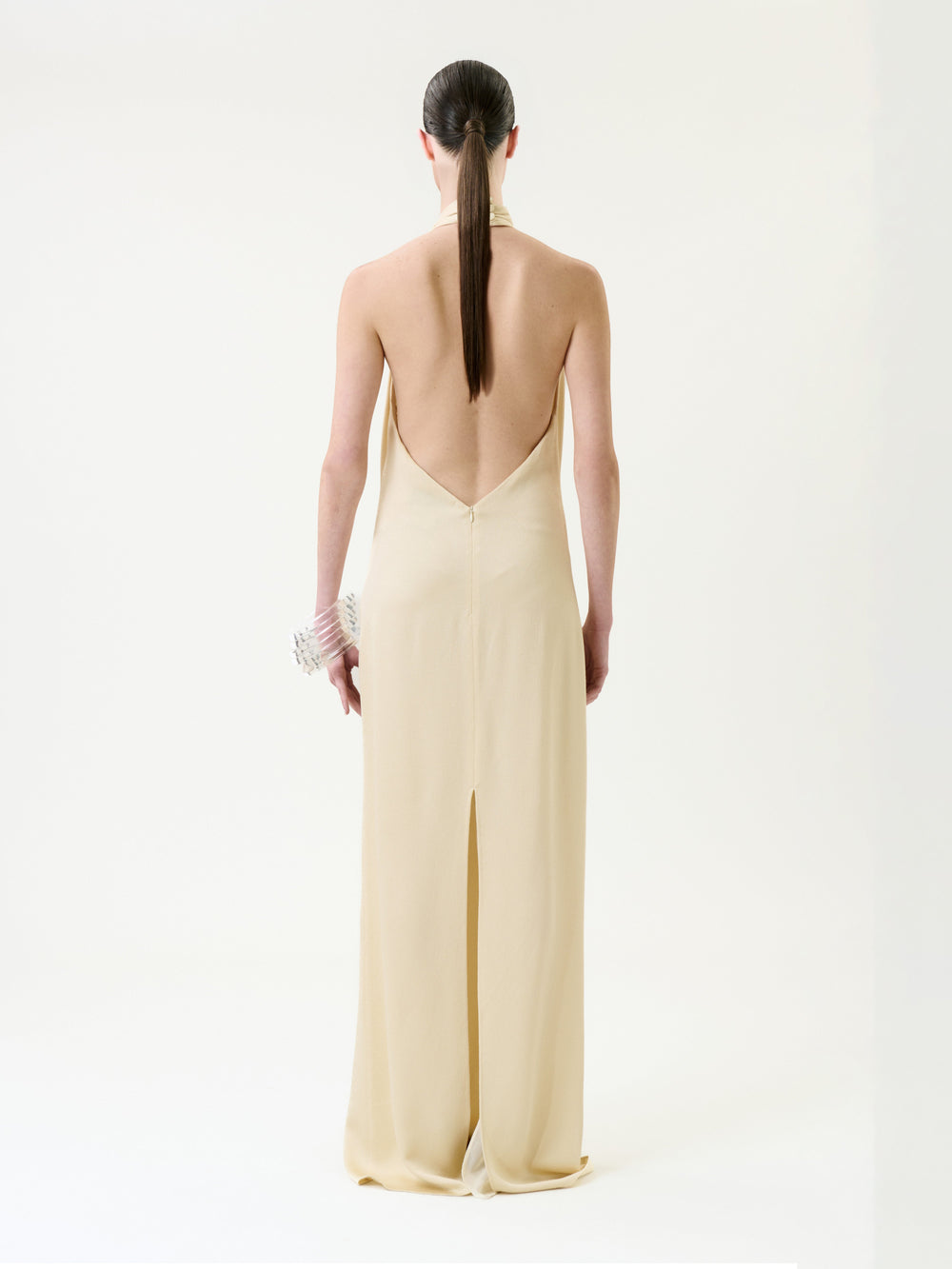 A person stands with their back to the camera, wearing the Maica Dress—a beige, floor-length design with a backless cut and center slit. Their hair is slicked into a low ponytail, and they hold a beaded accessory in their left hand against a plain white background.