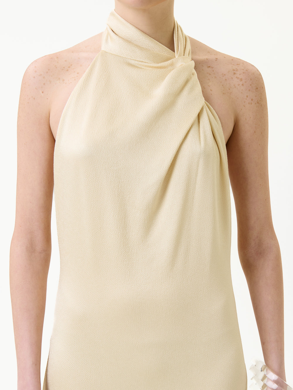 A person wearing the Maica Dress—a sleeveless, beige, halter-neck style with draped fabric—standing against a plain light background.