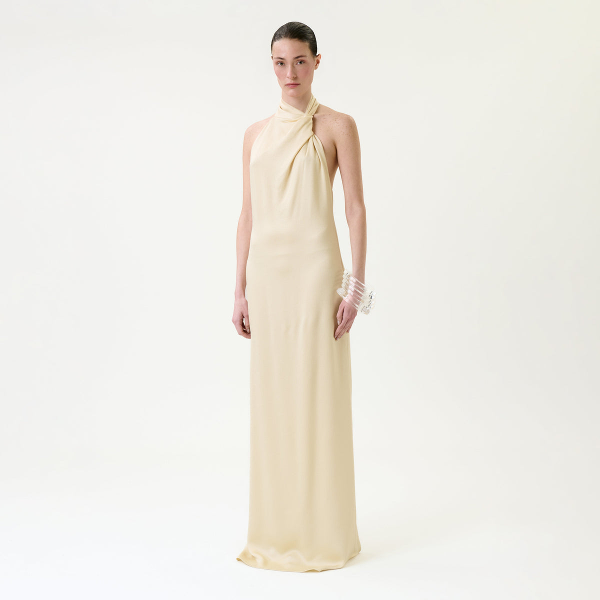 A person stands against a plain white background wearing the Maica Dress, a long, sleeveless, cream-colored halter dress, paired with a transparent bangle on their right wrist.